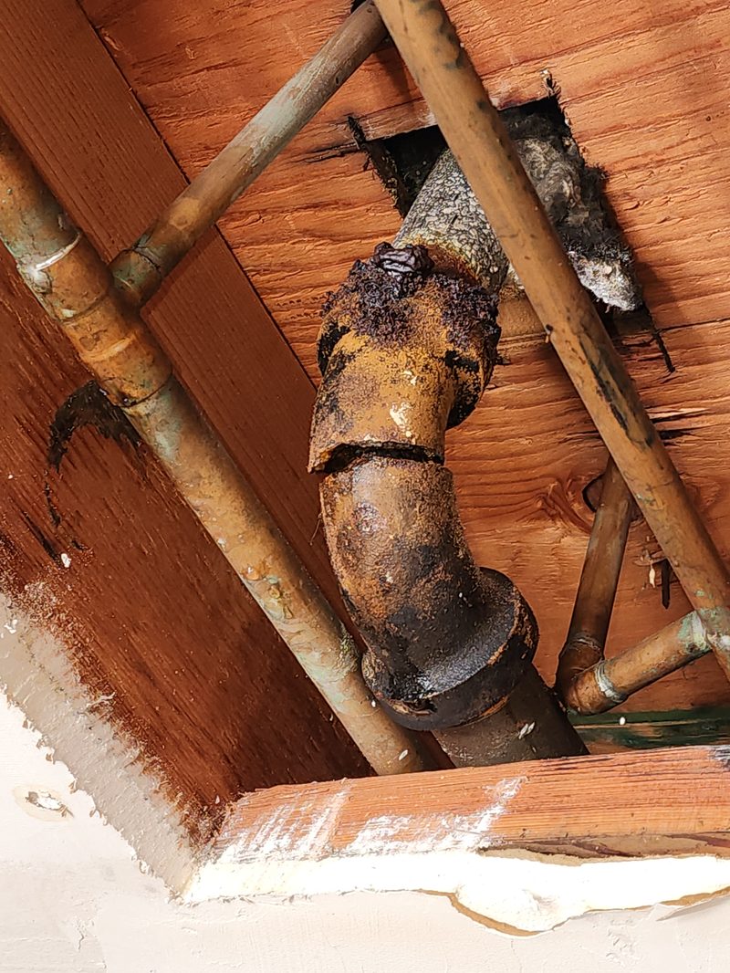 drainage-service-broken-cast-iron-elbow-orange-county Corroded cast iron drain and vent piping with aging copper lines under a house, seen against wood floor joists