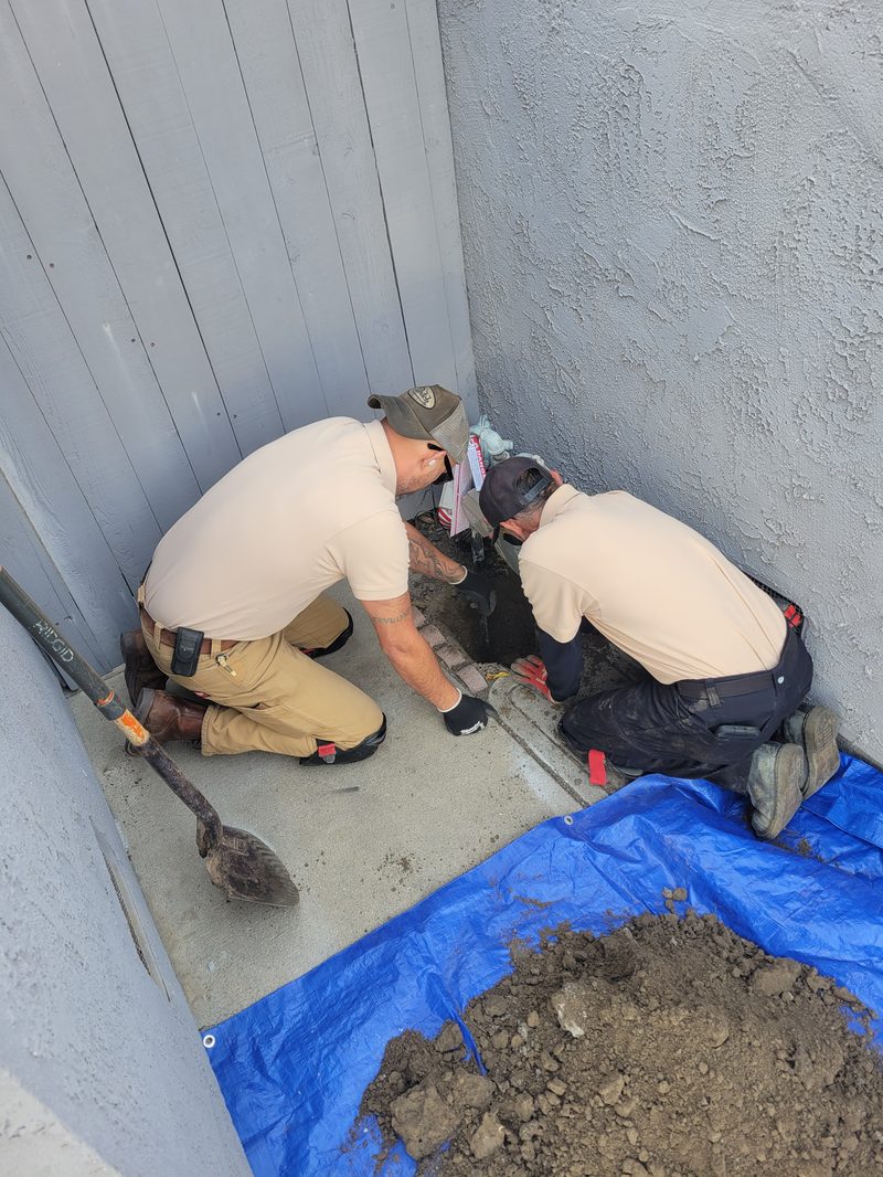 slab-leak-excavation-reroute-orange-county Two Professional Plumbing technicians excavating at the foundation to access and reroute a slab leak, soil piled on a blue tarp