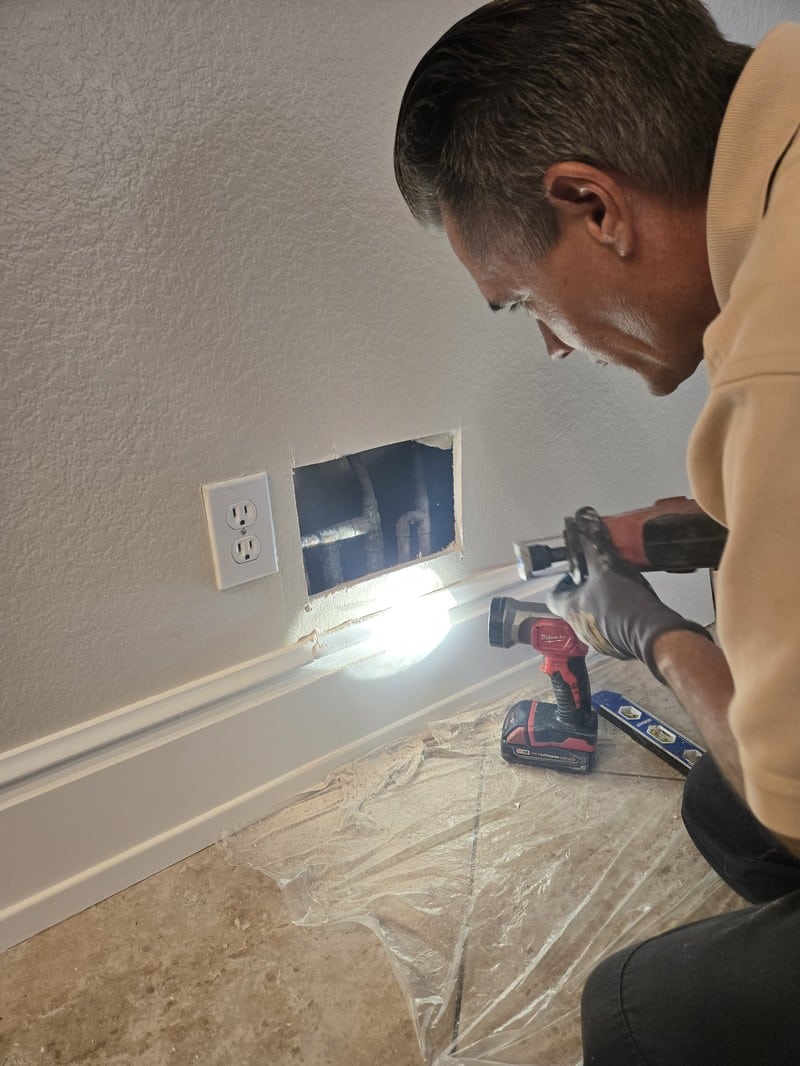 slab-leak-locating-with-flashlight-orange-county Plumber using a flashlight to inspect copper piping through an opening cut in the wall while pinpointing a slab leak
